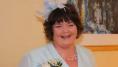 The late Alison McKinney - a woman with short, dark, hair - is smiling into the camera. She is dressed to attend a wedding and is wearing a pale blue jacket with a pin-on corsage of white flowers, a blue feather fascinator in her hair and a pearl-type necklace.
