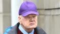 George Hendry. He is wearing a purple hat, light blue and grey raincoat, dark red jumper with white shirt underneath. A blurred wall is behind him.